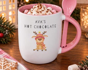 May include: A white ceramic mug with a pink interior and a matching pink spoon tucked into the handle. The mug features the text "AVA'S HOT CHOCOLATE" above a graphic of a reindeer wearing a pink and yellow striped hat and scarf, filled with whipped cream and marshmallows.