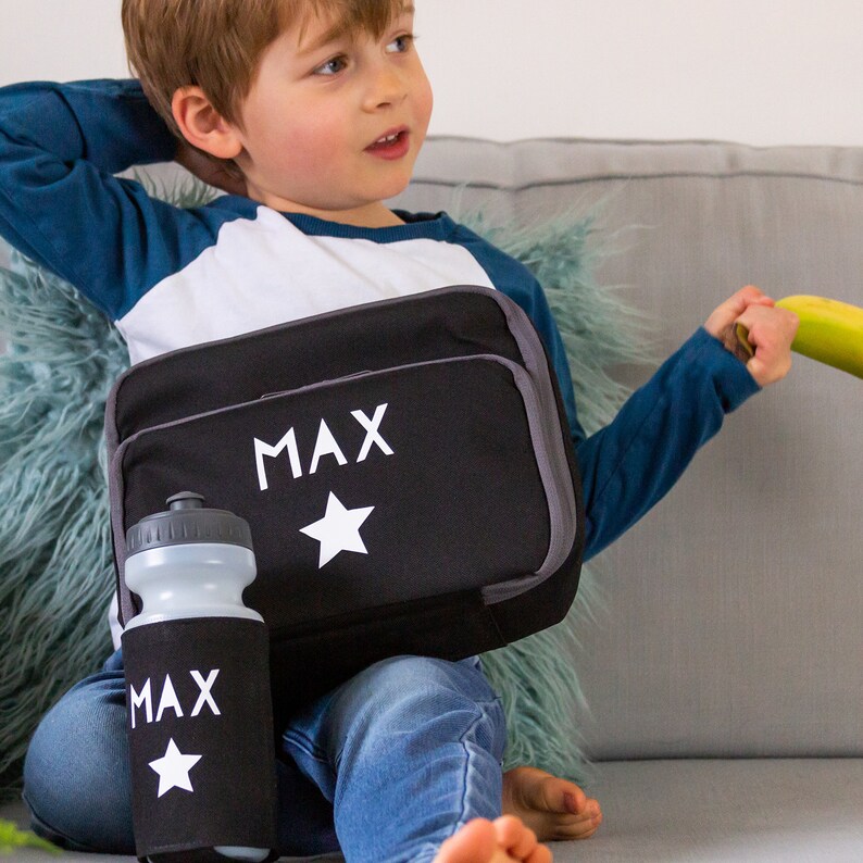 May include: Black lunch bag and water bottle set with the name "MAX" and a white star. The lunch bag has a gray trim and a matching water bottle. The water bottle is in a black sleeve with the name "MAX" and a white star.