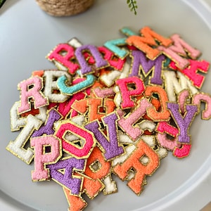 May include: A pile of colourful felt letter patches with gold glitter trim. The letters are in various colours, including pink, purple, orange, and white.