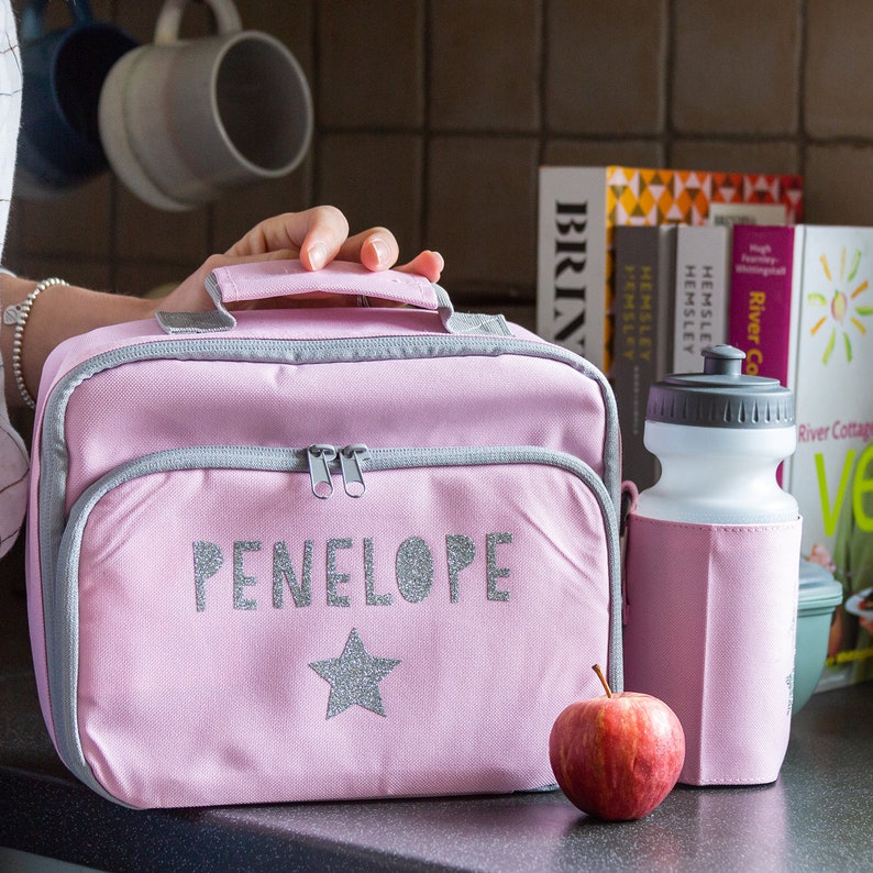 May include: Pink lunch bag with the name "PENELOPE" in silver glitter letters and a matching star. The bag has a gray trim and a handle. A water bottle with a pink sleeve and a red apple are next to the bag.