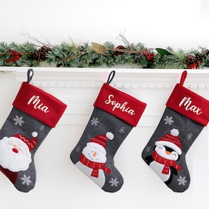 May include: Three personalised Christmas stockings with red cuffs and grey bodies. Each stocking features a different festive character: Santa Claus, a snowman, and a penguin. The names "Mia", "Sophia", and "Max" are embroidered on the cuffs.