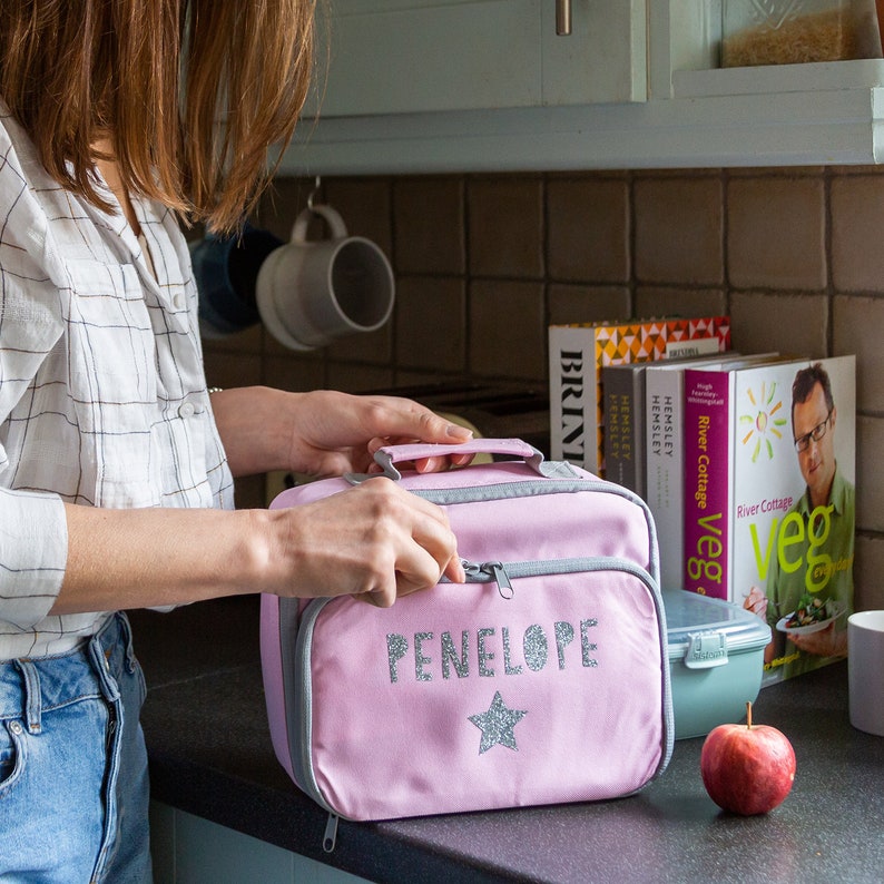 May include: A pink lunch bag with a gray trim and handle. The name "PENELOPE" is printed in silver glitter above a glitter star. The bag is on a black countertop next to a red apple and a green container.