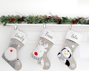 May include: Three gray Christmas stockings with white felt trim and personalized names: Emily, Finn, and Isla. Each stocking features a different embroidered character: Santa, a reindeer, and a penguin.