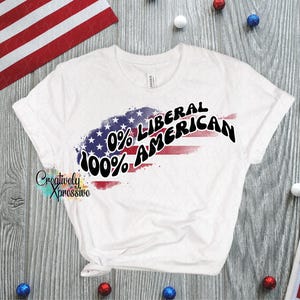 May include: White t-shirt with a patriotic design. The shirt features a stylized American flag with the text "0% Liberal 100% American" in black. The shirt is displayed on a wooden surface with decorative patriotic elements.