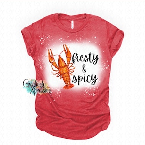 Puede incluir: Camiseta roja con una ilustración de acuarela de un cangrejo de río y las palabras "Fiesty & Spicy" en escritura negra. La camiseta tiene un diseño salpicado blanco alrededor del cangrejo de río y el texto. La marca "Creatively Xpressive" está en la esquina inferior izquierda.