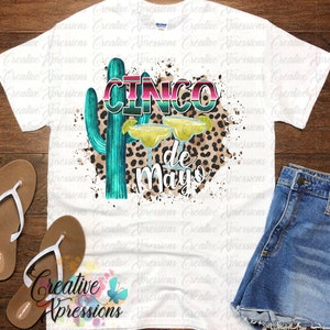 Pu&ograve; includere: Una maglietta bianca con un design grafico di un cactus, due bicchieri di margarita e il testo "Cinco de Mayo" in un font colorato e stratificato. Lo sfondo &egrave; un motivo di schizzi bianchi con una sovrapposizione di stampa leopardata.
