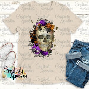 May include: A white t-shirt featuring a skull design surrounded by purple and orange flowers. The skull is detailed with black eyes and a white smile. The background is a light beige colour.