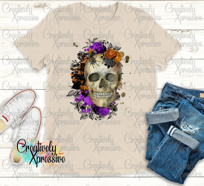 May include: A white t-shirt featuring a skull design surrounded by purple and orange flowers. The skull is detailed with black eyes and a white smile. The background is a light beige colour.