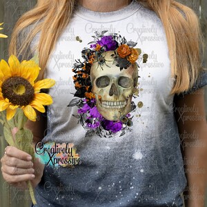 May include: A white t-shirt with a bleached effect featuring a skull with floral accents. The skull is surrounded by purple, orange, and black flowers. The text "Creatively Xpressivo" is printed on the shirt.