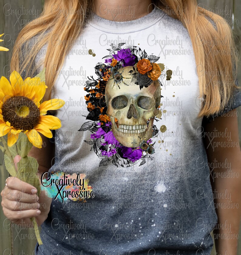May include: A white t-shirt with a bleached effect featuring a skull with floral accents. The skull is surrounded by purple, orange, and black flowers. The text "Creatively Xpressivo" is printed on the shirt.