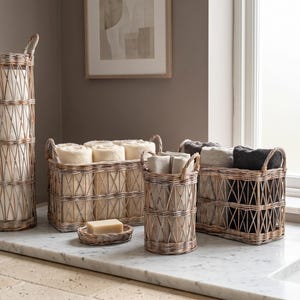 Può includere: Una collezione di cesti portaoggetti in vimini intrecciato di varie forme e dimensioni, tra cui un cesto cilindrico alto, un cesto rettangolare e un piccolo portasapone ovale, contenenti asciugamani piegati color crema, beige e grigio scuro.