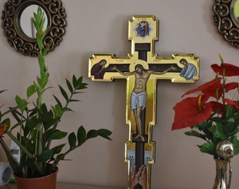 May include: A gold-toned wooden cross with a depiction of Jesus Christ on the cross. The cross is decorated with intricate details and has a small, ornate frame around the image of Jesus. The cross is displayed on a wooden shelf with a potted plant and a decorative vase.