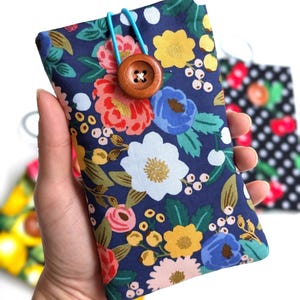 Puede incluir: Una funda para teléfono azul con estampado floral, cierre de botón de madera y correa elástica turquesa.