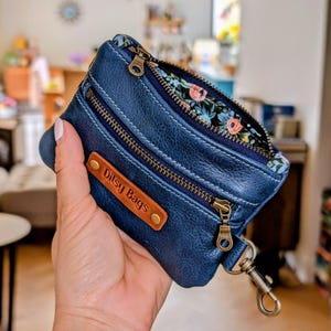 May include: A navy blue leather wallet with three zipped compartments and a floral patterned interior. The wallet has a small brown leather tag that reads "Ditsy Bags". A silver-coloured clasp is attached to the wallet.