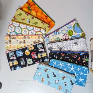 May include: A collection of ten zippered pouches with various colourful patterns. The pouches feature designs such as a periodic table, cameras, bicycles, cats, and a spider web.