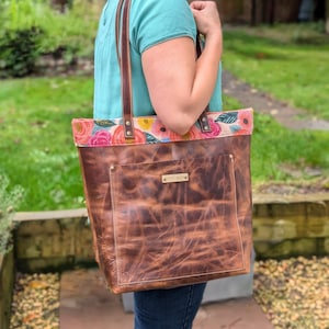May include: A brown leather tote bag with a floral fabric lining. The bag has a front pocket and a leather strap with a metal tag that says "Monterey Bag".