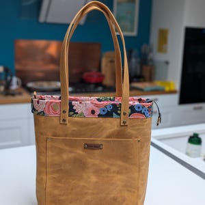 May include: A tan tote bag with a floral patterned top and handles. The bag has a front pocket and a small metal tag. The interior lining features a colorful floral design with pink and blue flowers.