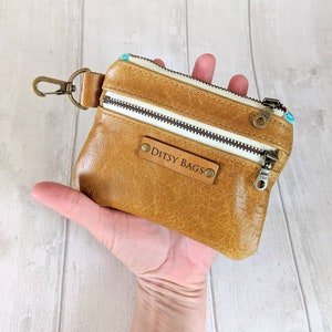 May include: A tan leather coin purse with two zippered compartments and a key ring clasp. The purse has a label that reads "Ditsy Bags". The zippers are white, and the interior fabric has a floral pattern.