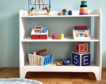 May include: White wooden bookshelf with three shelves, filled with colorful wooden toys, including a toy cash register, a toy scale, a toy shopping basket, a toy phone, a toy abacus, and a toy stacking ring tower.