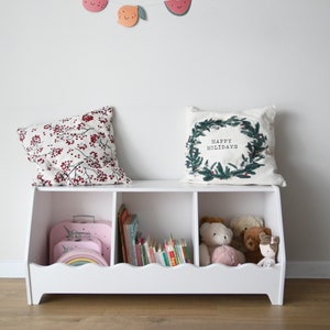 May include: White wooden storage bench with three compartments. The top compartment is empty, the middle compartment is filled with books, and the right compartment is filled with stuffed animals. A pillow with the text "Happy Holidays" is on top of the bench. A fruit garland with a watermelon, apple, pear, banana, and strawberry hangs on the wall behind the bench.