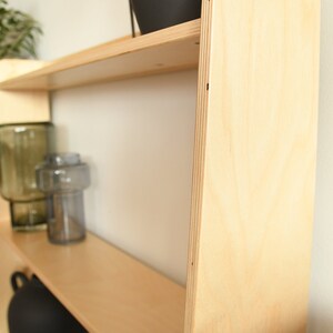 May include: A light wood shelf unit with rounded edges and two shelves. The shelves are empty, but there are decorative items on the top shelf, including a black planter and two glass vases.