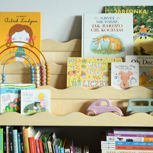 May include: A wooden bookshelf with multiple shelves, filled with a variety of children's books. The top shelf has a rainbow-colored abacus with wooden beads. The bottom shelf has a collection of hardcover books with black covers. The middle shelf has a pink and a blue toy car.