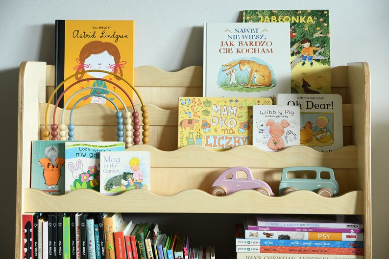 May include: A wooden bookshelf with multiple shelves, filled with a variety of children's books. The top shelf has a rainbow-colored abacus with wooden beads. The bottom shelf has a collection of hardcover books with black covers. The middle shelf has a pink and a blue toy car.