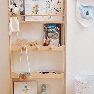 May include: A wooden bookshelf with five shelves, featuring a variety of children's books, toys, and decorations. The top shelf has three books, including one with the title "O zimie" and another with the title "Szcze cie". The second shelf has a book titled "W Arktyce" and a brown teddy bear. The third shelf has a wooden toy camera and a wooden toy stacking ring. The fourth shelf has a book titled "Naciśnij mnie". The bottom shelf is empty. The bookshelf is against a white wall with a white lamp and a koala-shaped wall hanging.