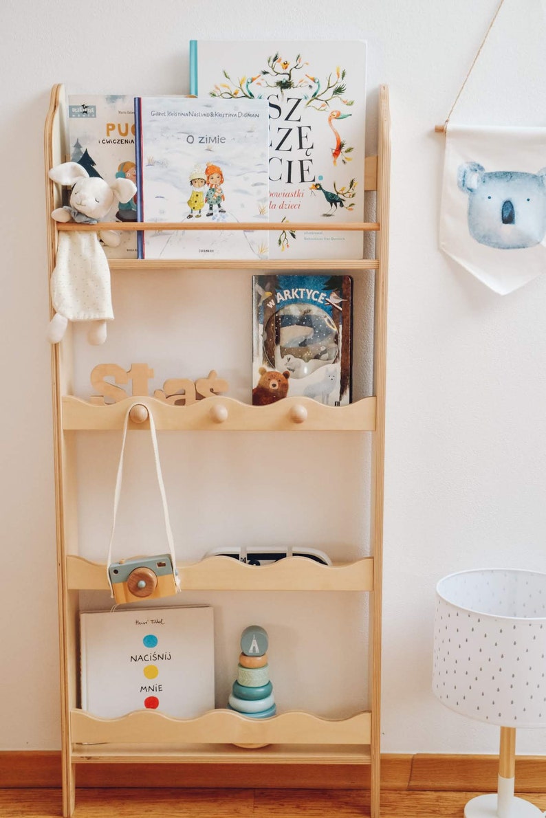 May include: A wooden bookshelf with five shelves, featuring a variety of children's books, toys, and decorations. The top shelf has three books, including one with the title "O zimie" and another with the title "Szcze cie". The second shelf has a book titled "W Arktyce" and a brown teddy bear. The third shelf has a wooden toy camera and a wooden toy stacking ring. The fourth shelf has a book titled "Naciśnij mnie". The bottom shelf is empty. The bookshelf is against a white wall with a white lamp and a koala-shaped wall hanging.