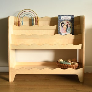May include: A light wood bookshelf with wavy edges on the shelves. The bookshelf has two shelves and is filled with books and toys. The top shelf has a rainbow-colored abacus and a book titled "Small Kobliiki". The bottom shelf has a wicker basket filled with colorful wooden blocks.