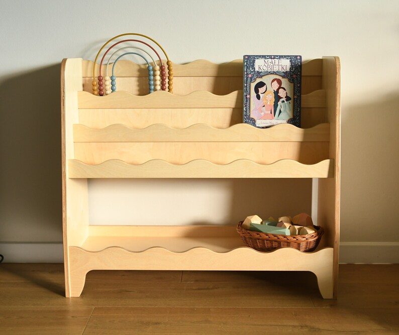 May include: A light wood bookshelf with wavy edges on the shelves. The bookshelf has two shelves and is filled with books and toys. The top shelf has a rainbow-colored abacus and a book titled "Small Kobliiki". The bottom shelf has a wicker basket filled with colorful wooden blocks.