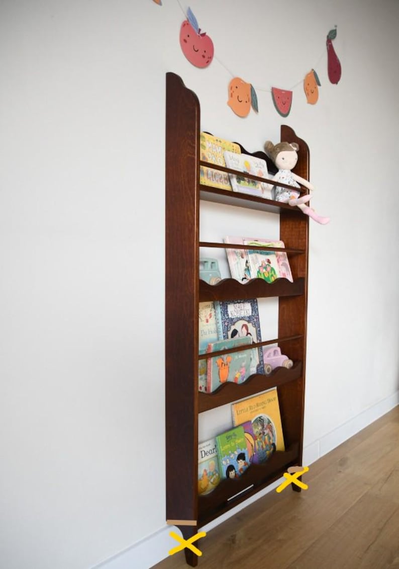 May include: A tall, dark wooden bookshelf with multiple shelves, filled with children's books and a doll. A garland of colorful fruit decorations hangs above. The bookshelf is approximately 60 inches tall.