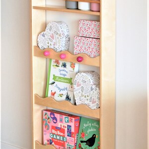 May include: A wooden shelf unit with five shelves, featuring a variety of items including books, boxes, and potted plants. The shelf unit is painted in a light wood color and has a decorative scalloped edge. The shelf unit is standing on a wooden floor.