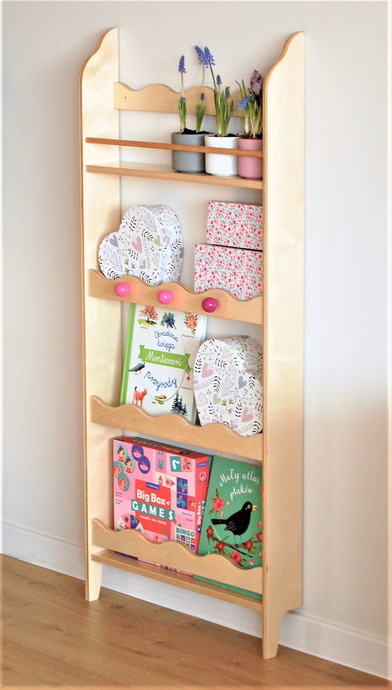 May include: A wooden shelf unit with five shelves, featuring a variety of items including books, boxes, and potted plants. The shelf unit is painted in a light wood color and has a decorative scalloped edge. The shelf unit is standing on a wooden floor.