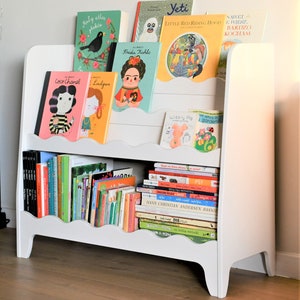 May include: White bookshelf with two shelves, featuring a variety of colorful children's books. The top shelf has books with titles like "Frida Kahlo", "Yeti", "Little Red Riding Hood", and "Coco Chanel". The bottom shelf has a stack of books with titles like "Hans Christian Andersen Basnie", "PSY", and "Wibbly Pig".