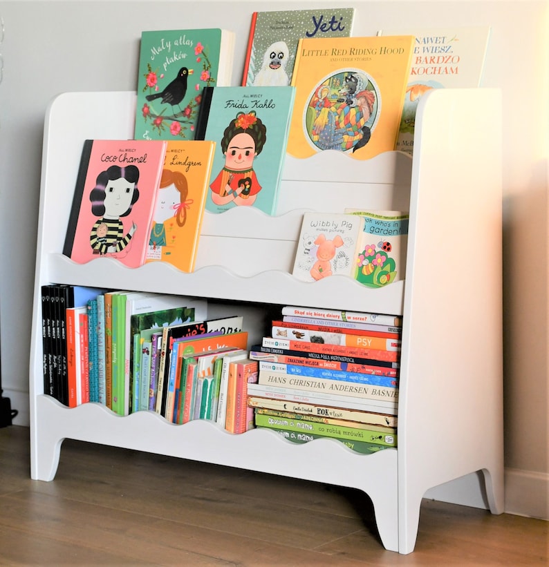 May include: White bookshelf with two shelves, featuring a variety of colorful children's books. The top shelf has books with titles like "Frida Kahlo", "Yeti", "Little Red Riding Hood", and "Coco Chanel". The bottom shelf has a stack of books with titles like "Hans Christian Andersen Basnie", "PSY", and "Wibbly Pig".