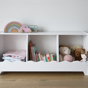 Könnte beinhalten: Weißer, hölzerner Spielzeug-Aufbewahrungsschrank mit drei Fächern. Das obere Fach hat ein Regenbogen-Holzspielzeug und ein rosa Spielzeugauto. Das mittlere Fach hat Bücher. Das untere Fach hat Stofftiere und eine Decke.
