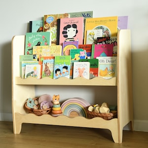 May include: A wooden bookshelf with two shelves, featuring a variety of colorful children's books. The bookshelf has a scalloped edge and is filled with books with titles like "Coco Chanel", "Little Red Riding Hood", and "Oh Dear!"