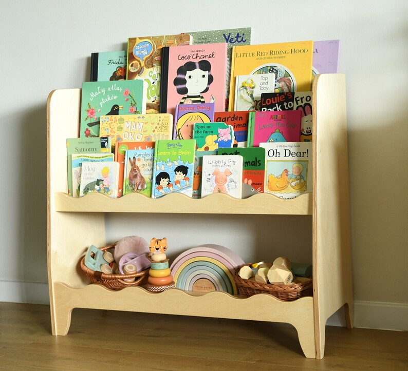 May include: A wooden bookshelf with two shelves, featuring a variety of colorful children's books. The bookshelf has a scalloped edge and is filled with books with titles like "Coco Chanel", "Little Red Riding Hood", and "Oh Dear!"