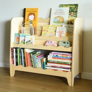 May include: A light wood bookshelf with rounded edges and shelves for storing books. The shelves are filled with a variety of children's books, including some with colorful illustrations. There are also two toy cars on the top shelf.