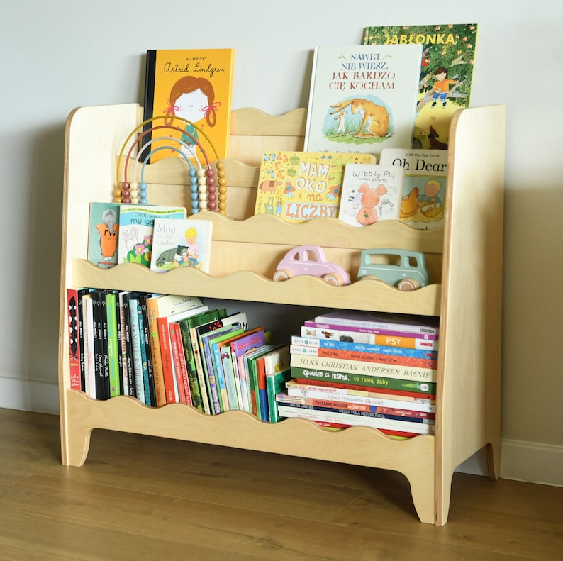 May include: A light wood bookshelf with rounded edges and shelves for storing books. The shelves are filled with a variety of children's books, including some with colorful illustrations. There are also two toy cars on the top shelf.