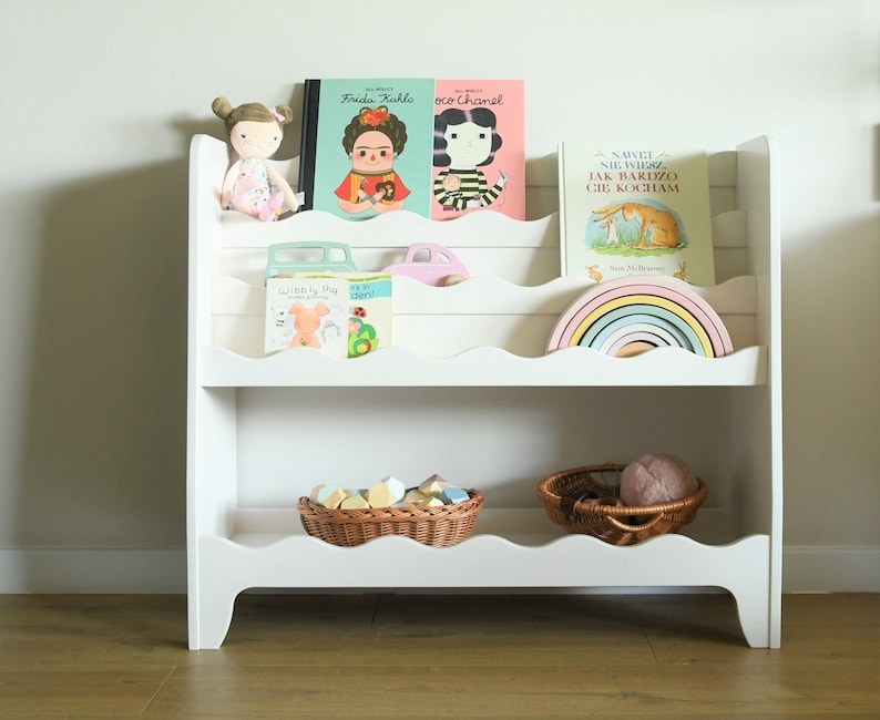 May include: White wooden bookshelf with five shelves, featuring a rainbow stacking toy, a doll, and several children's books. The books include titles like "Frida Kahlo", "Coco Chanel", and "Nawei Niewiesz Jak Bardzo Cie Kocham".