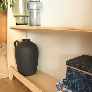 May include: A wooden shelf unit with three shelves. The top shelf has two glass vases, one clear and one green. The middle shelf has a black ceramic vase. The bottom shelf has two decorative boxes, one with a blue and black pattern and one with a floral pattern.