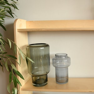 May include: Two glass vases on a wooden shelf. The vase on the left is a dark gray color and the vase on the right is a light gray color.