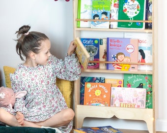 May include: A wooden bookshelf with multiple shelves filled with colorful children's books. The bookshelf has a light wood finish and is standing on a light wood floor. A young girl is sitting on a green cushion in front of the bookshelf, holding a book in her hands.