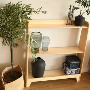 May include: A light wood bookshelf with three shelves. The top shelf has a metal sculpture and a black vase with a green plant. The middle shelf has two glass vases. The bottom shelf has a black vase and two boxes with floral patterns.