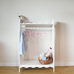 Op de afbeelding: Een wit houten kledingrek met een geschulpte boven- en onderrand. Het heeft een horizontale stang met roze kledinghangers in de vorm van wolken, een lichtblauw overhemd, een kleine pop op de plank en een rieten mand met knuffels onderin.