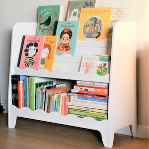 May include: White bookshelf with wavy edges and two shelves. The top shelf has colorful children's books with titles like "Frida Kahlo", "Yeti", "Little Red Riding Hood", and "Coco Chanel". The bottom shelf has a stack of books with titles like "Hans Christian Andersen Basnie", "PSY", and "Wibbly Pig".