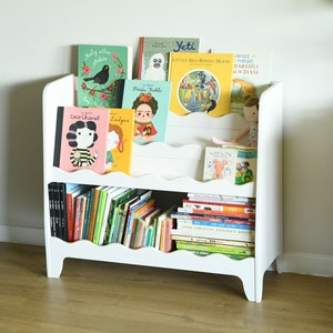 May include: White bookshelf with two shelves, filled with colorful children's books. The top shelf has a variety of books with titles like "Little Red Riding Hood" and "Frida Kahlo". The bottom shelf is filled with smaller books.