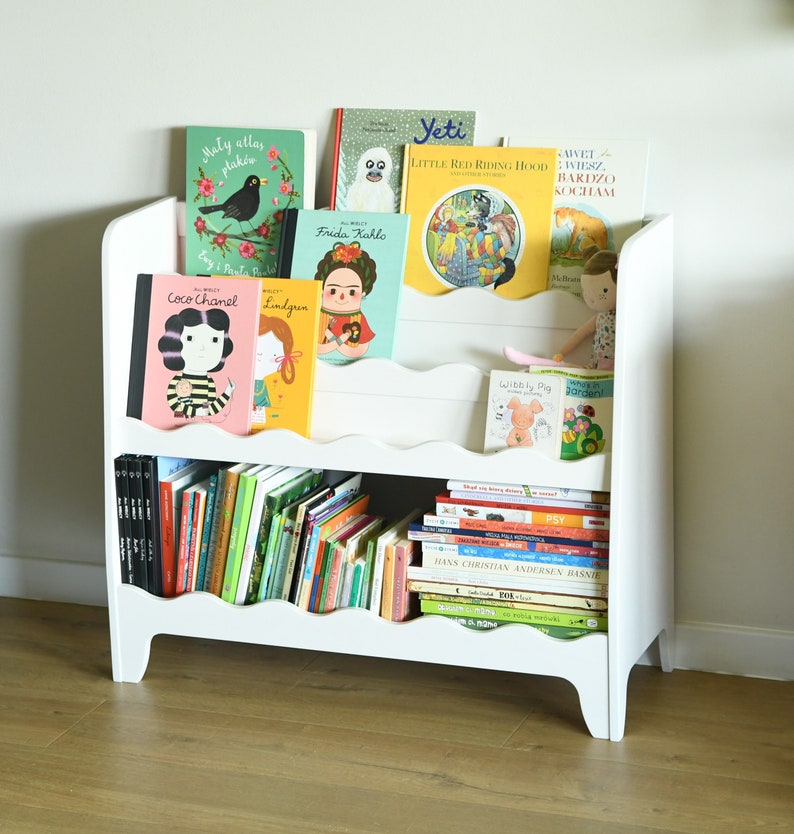 May include: White bookshelf with two shelves, filled with colorful children's books. The top shelf has a variety of books with titles like "Little Red Riding Hood" and "Frida Kahlo". The bottom shelf is filled with smaller books.